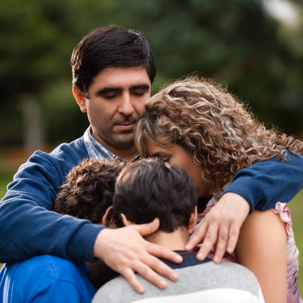 A man and woman hugging two boys closely outdoors with eyes closed and comforting expressions.