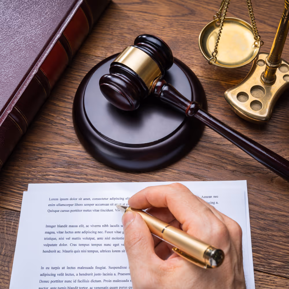 Hand holding gold pen writing on paper with judge's gavel, law book, and scales on wood table.