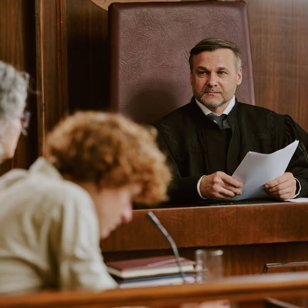 Judge in black robe holding papers seated behind courtroom bench, two people in foreground.