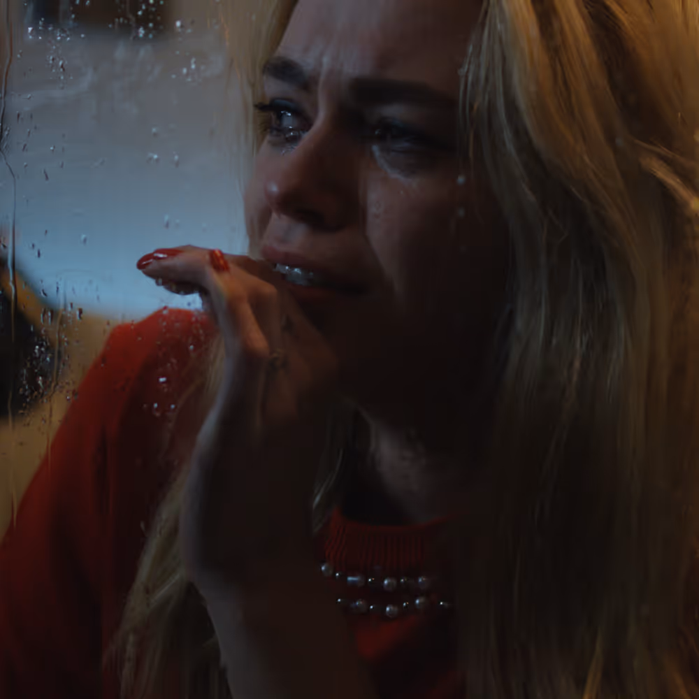 Woman with blonde hair crying and touching a rain-spotted window, wearing a red sweater with pearls.