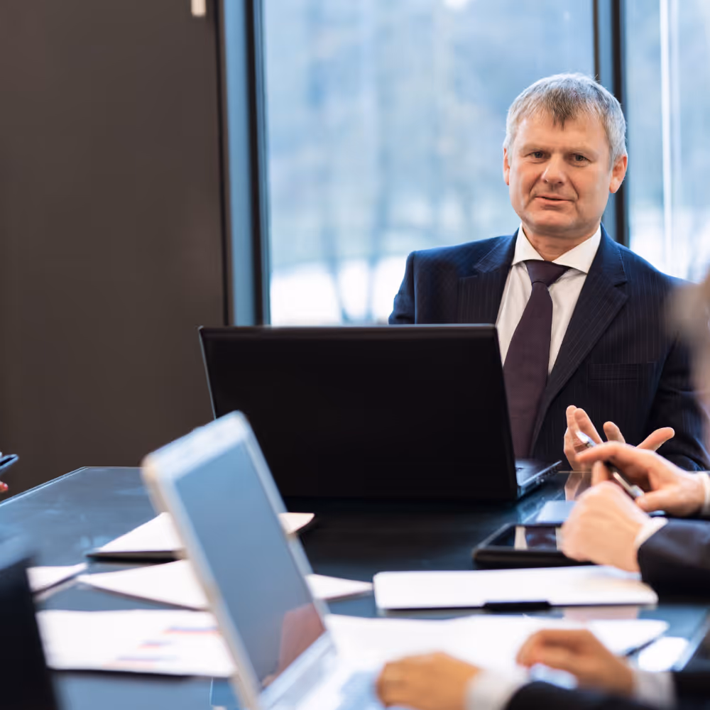 Businessman in suit speaking at a meeting around a table with laptops and papers.