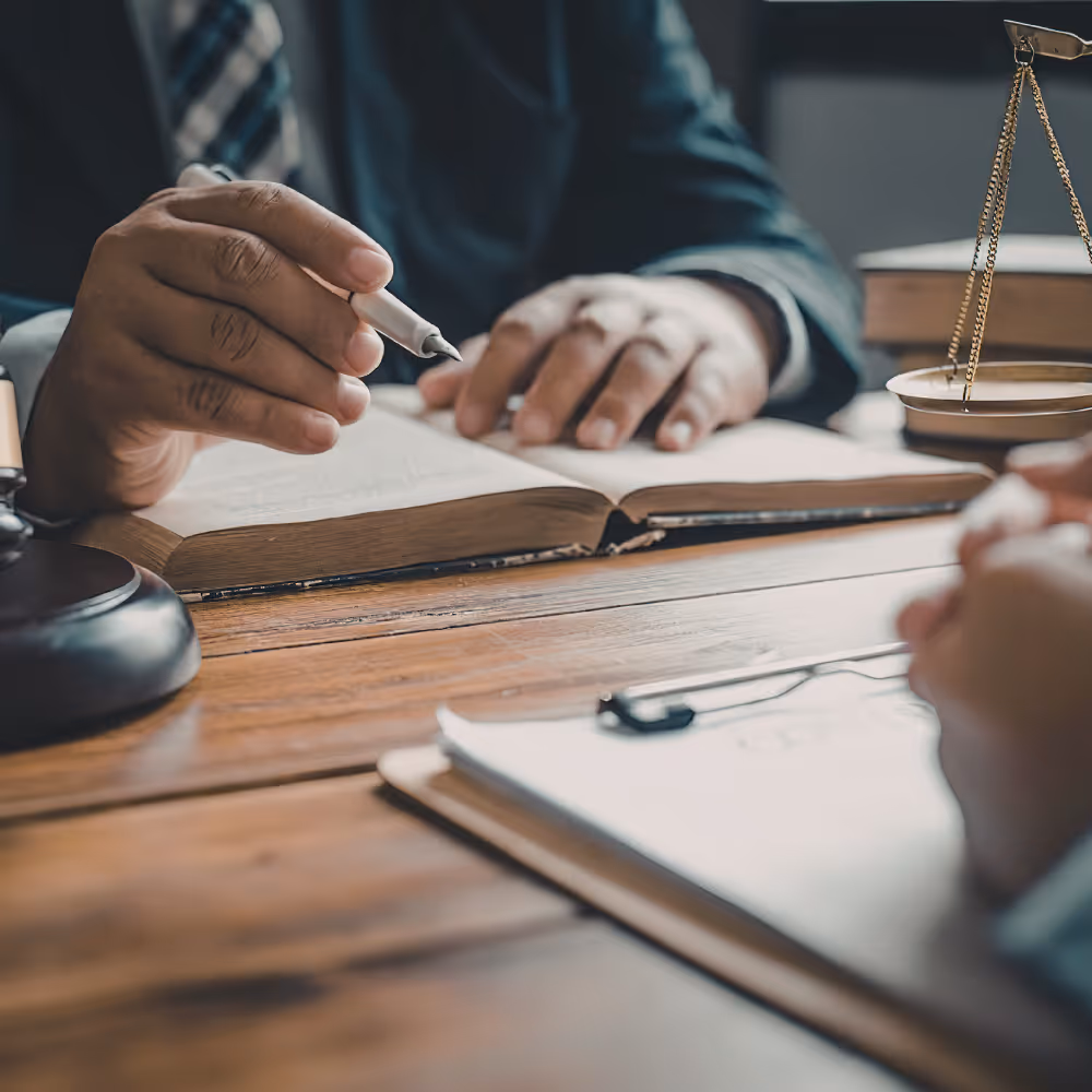 Person in suit holding pen over open book on wooden table with scales of justice nearby.