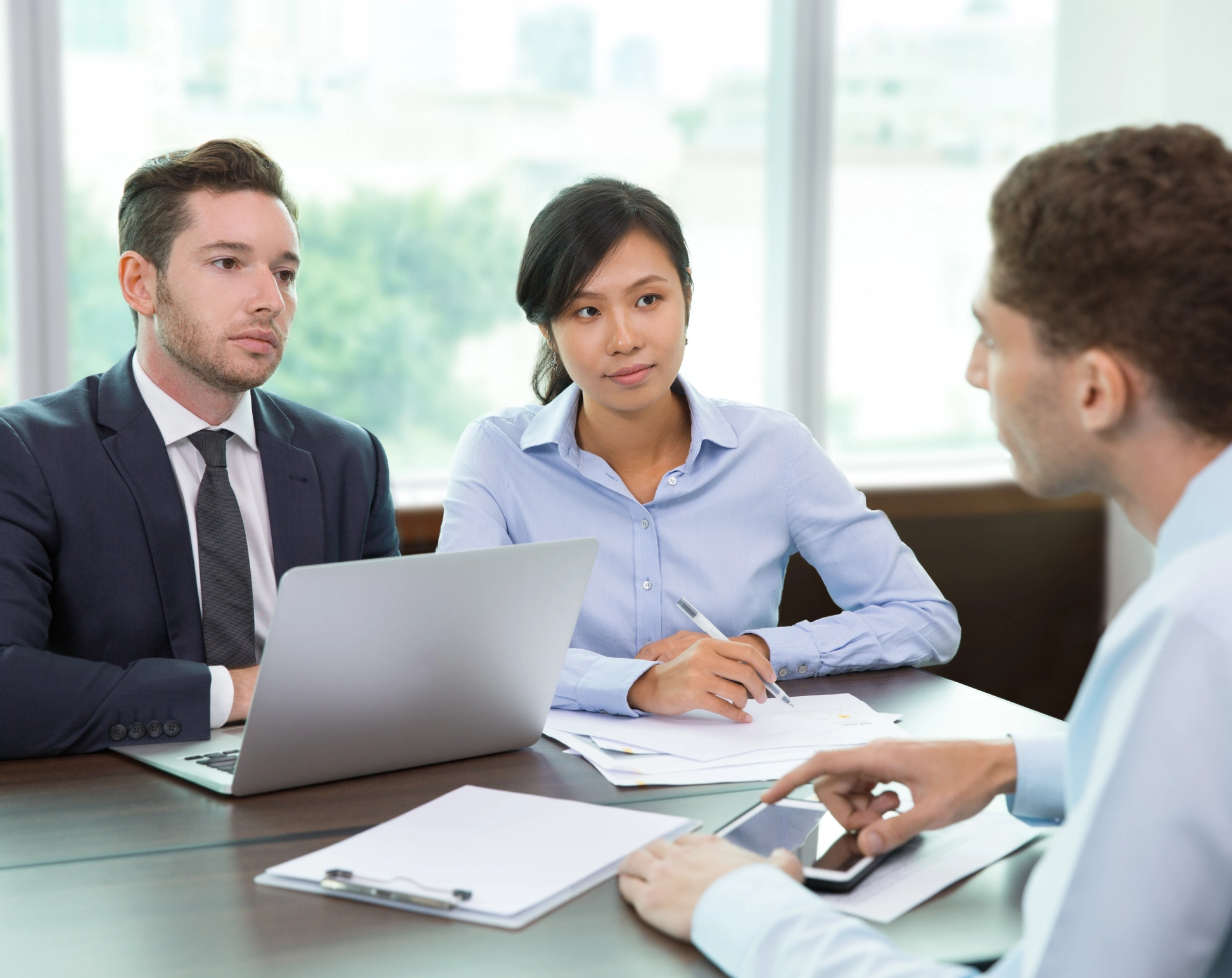 Three professionals in a meeting room with laptop, documents, and smartphone discussing serious matters.